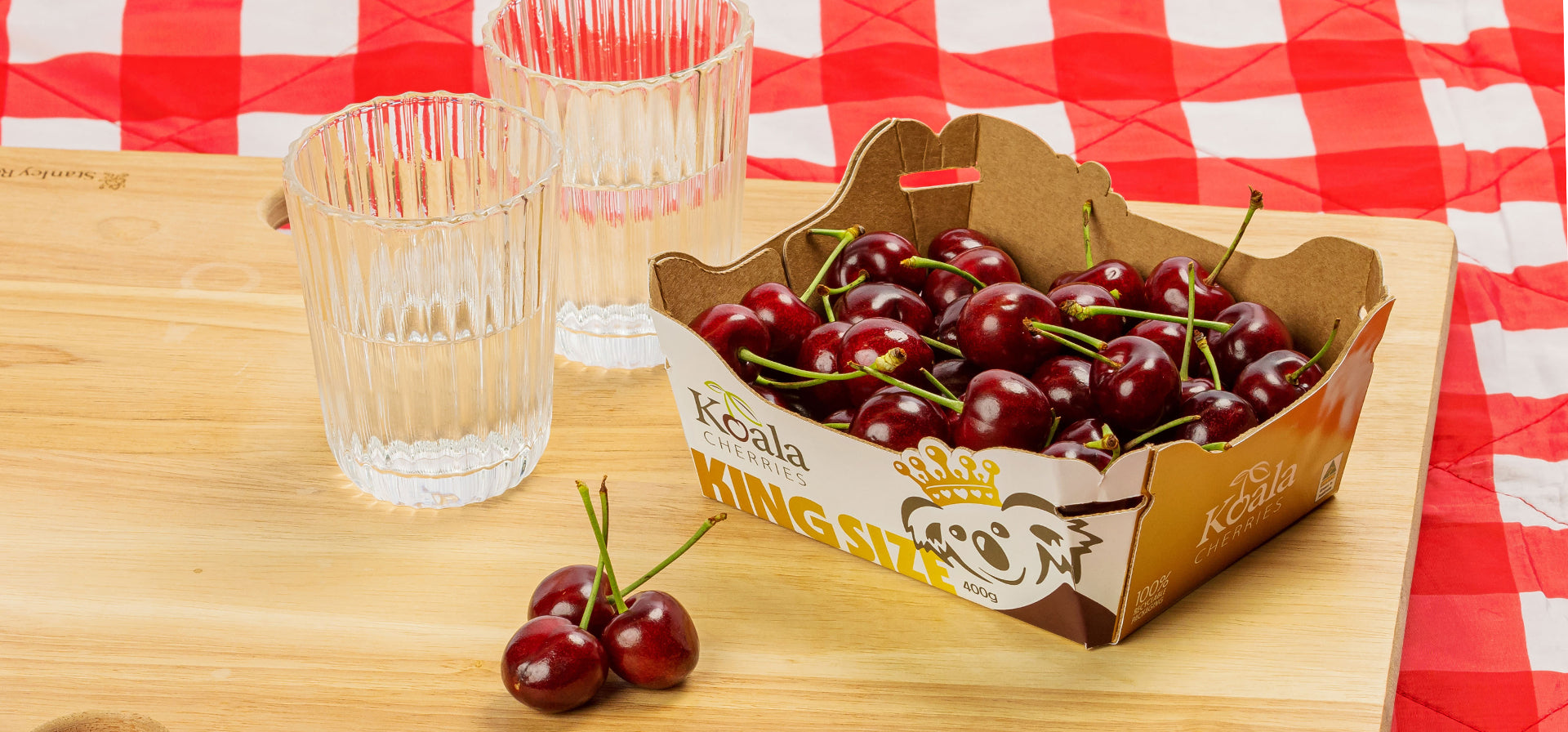 Koala king size cherries in cardboard recyclable packaging on a picnic rug with a wooden chopping board and two glasses of water